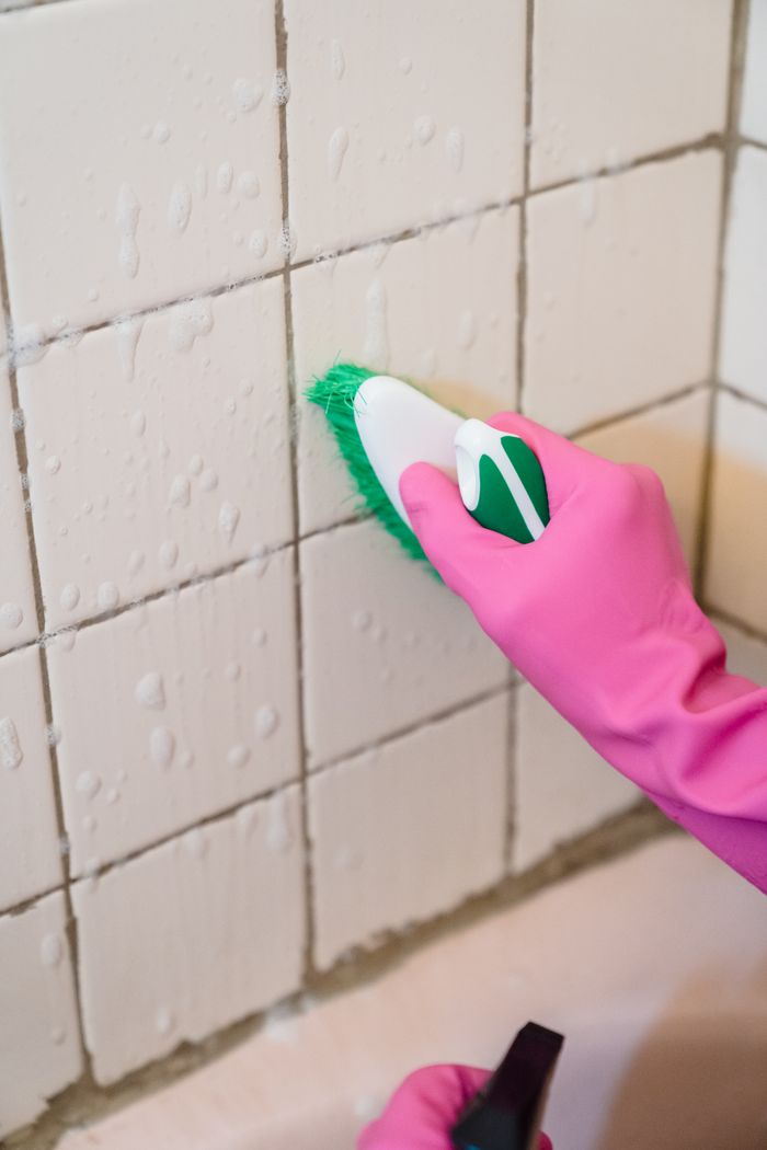 project-01-b Une main gantée de rose frotte des carreaux de salle de bain avec une brosse verte pour enlever la saleté.